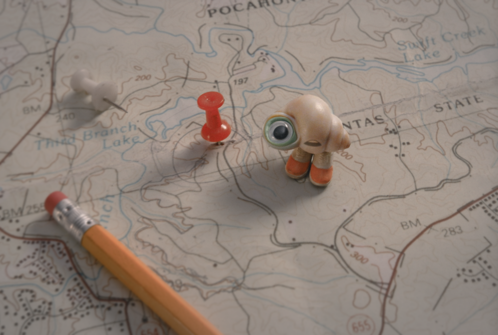 A shell with one eye is sitting on a map with a pencil and a thumbtack on the surface.