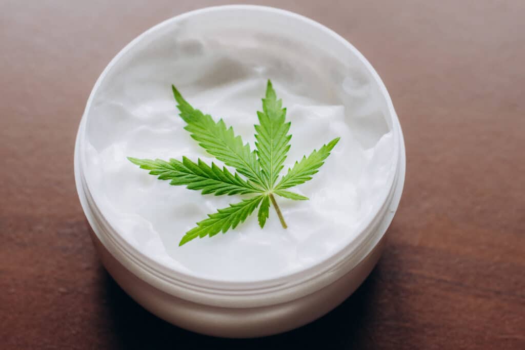 A bowl of moisturiser sits on a wooden background. A single cannabis leaf sits on the top of the open product.