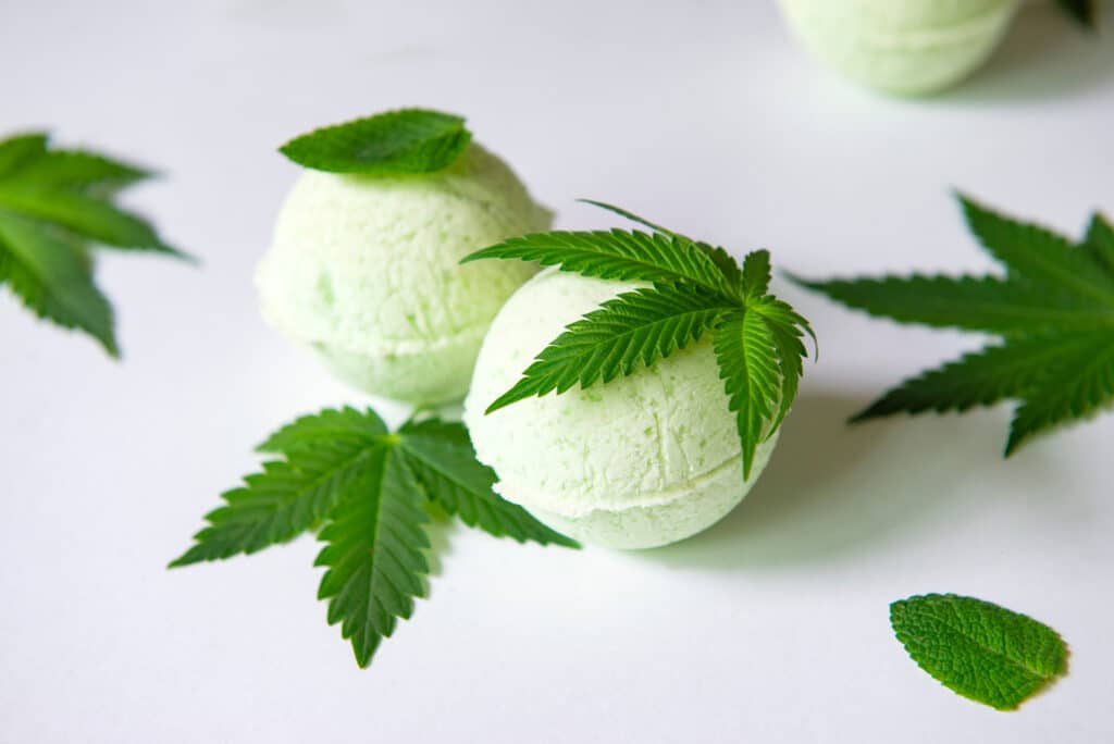 Two green bath bombs sitting on a white background. Cannabis leaves are scattered around and on top of them.
