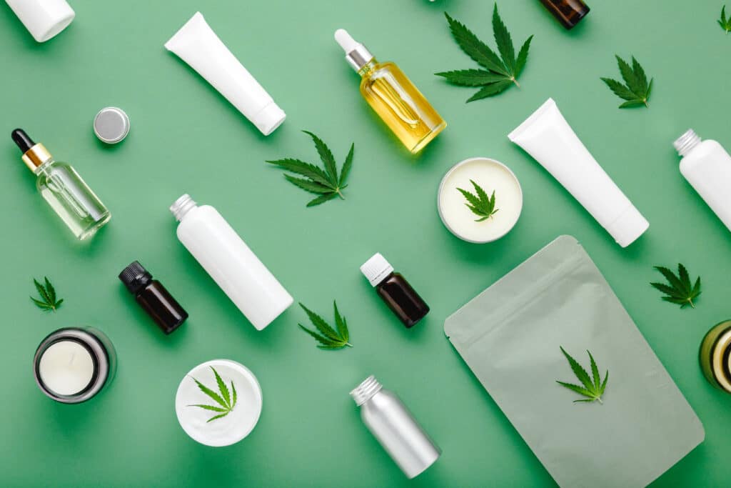 Various oils, creams, and blank skincare product bottles are laid out on a green background. Cannabis leaves are scattered on top of the products.