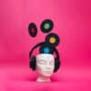 A mannequin head wearing headphones with vinyl records emerging from the top of the head. Pink background. Contemporary art with a retro music concept.
