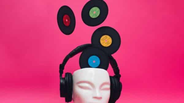 A mannequin head wearing headphones with vinyl records emerging from the top of the head. Pink background. Contemporary art with a retro music concept.