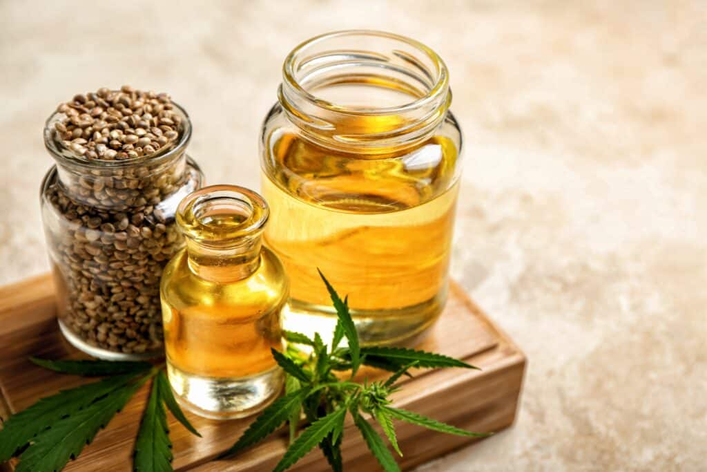 Three glass bottles sit on a wooden board. One is filled with Hemp seeds, while the other two are filled with yellow oil. Cannabis leaves sit underneath the glasses.