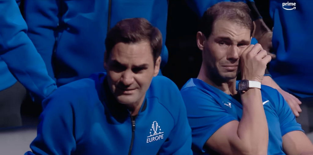 Roger Federer and Rafael Nadal sat beside each other. Two men, emotional, sat beside eac other. Dressed in blue. One is wiping his eyes.