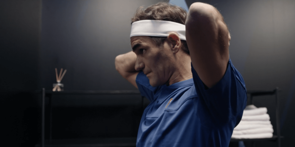 Roger Federer tying his headband. A man against a black background, dimly lit. He ties a white bandana around his head.