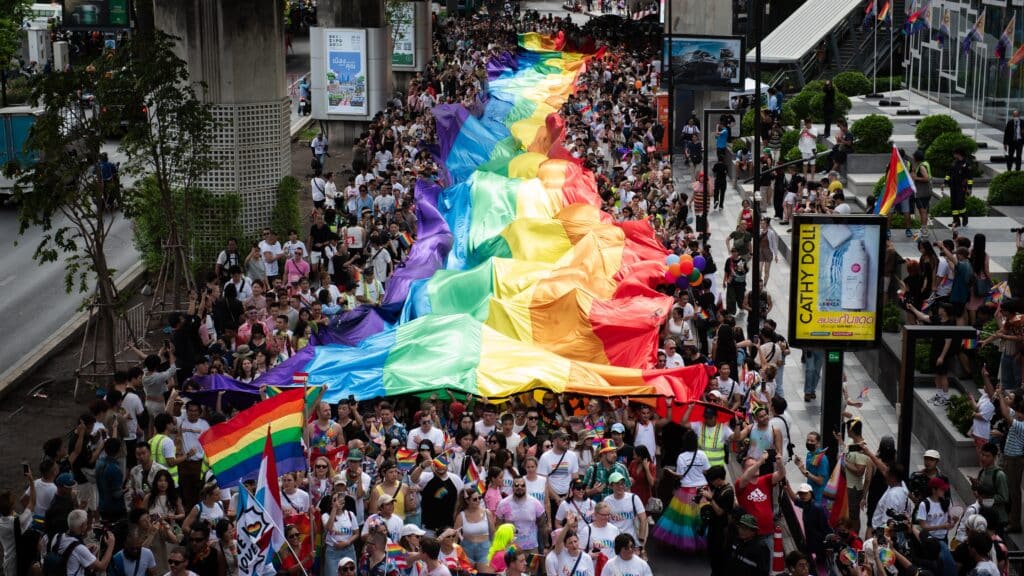 Massive Pride flag is unfurled above a crowd of people walking through Thailand.