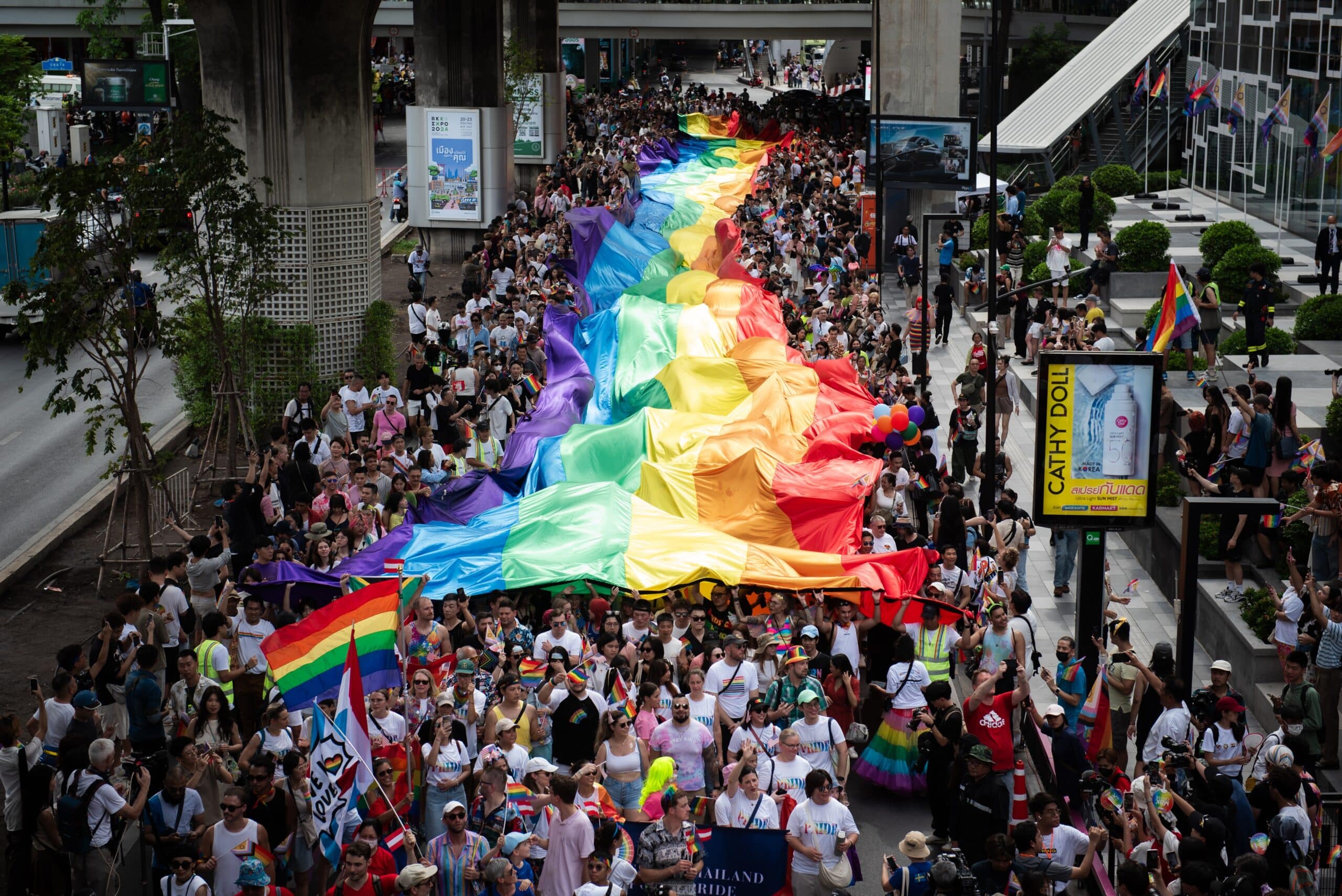 Massive Pride flag is unfurled above a crowd of people walking through Thailand.