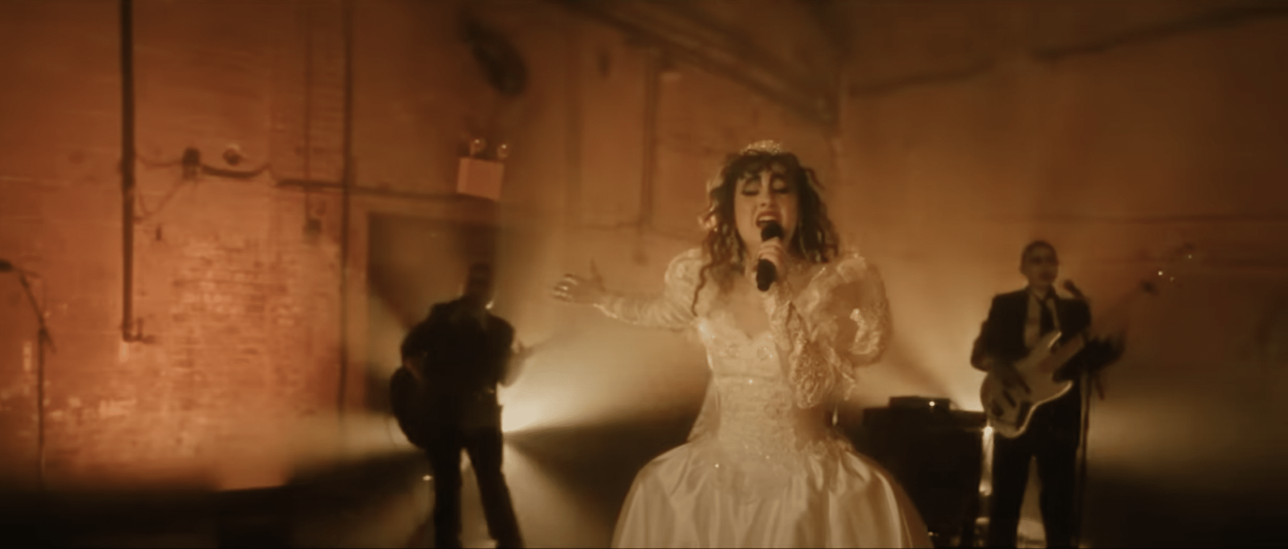 Chappell Roan wears a wedding dress in an industrial set. Light shines behind her as she holds a microphone. Behind her are two guitarists.