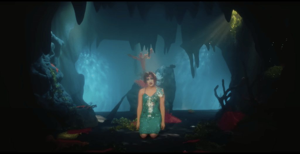 Chappell Roan sits on her knees in a teal sequin dress. She is in the foreground of a themed underwater cave set.