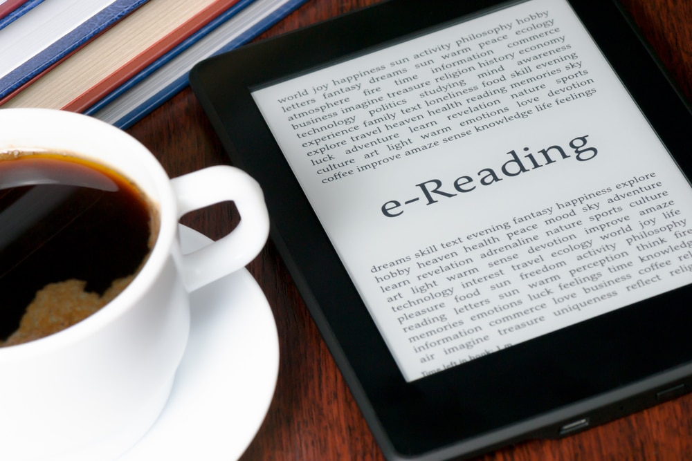 photo of an e-reader on the table next to a cup and saucer filled with black coffee. The e-reader has generated text at the top and bottom of the screen, in the middle in large writing is the word e-Reader to show an alternative way of reading sustainably