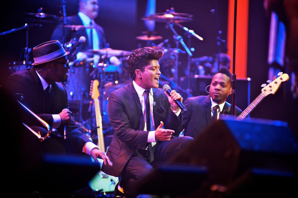 Bruno Mars is on stage kneeling in front of a monitor. Two of his bandmates are next to him.