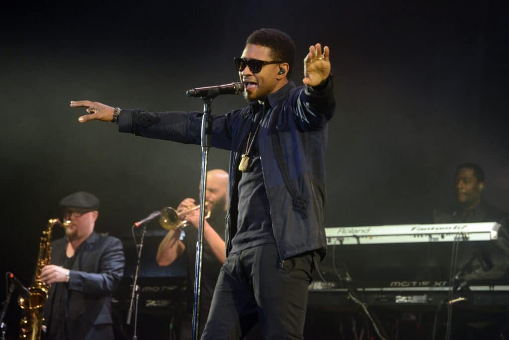 Usher is performing on a stage wearing sunglasses. He has his hands stretched out diagonally. 