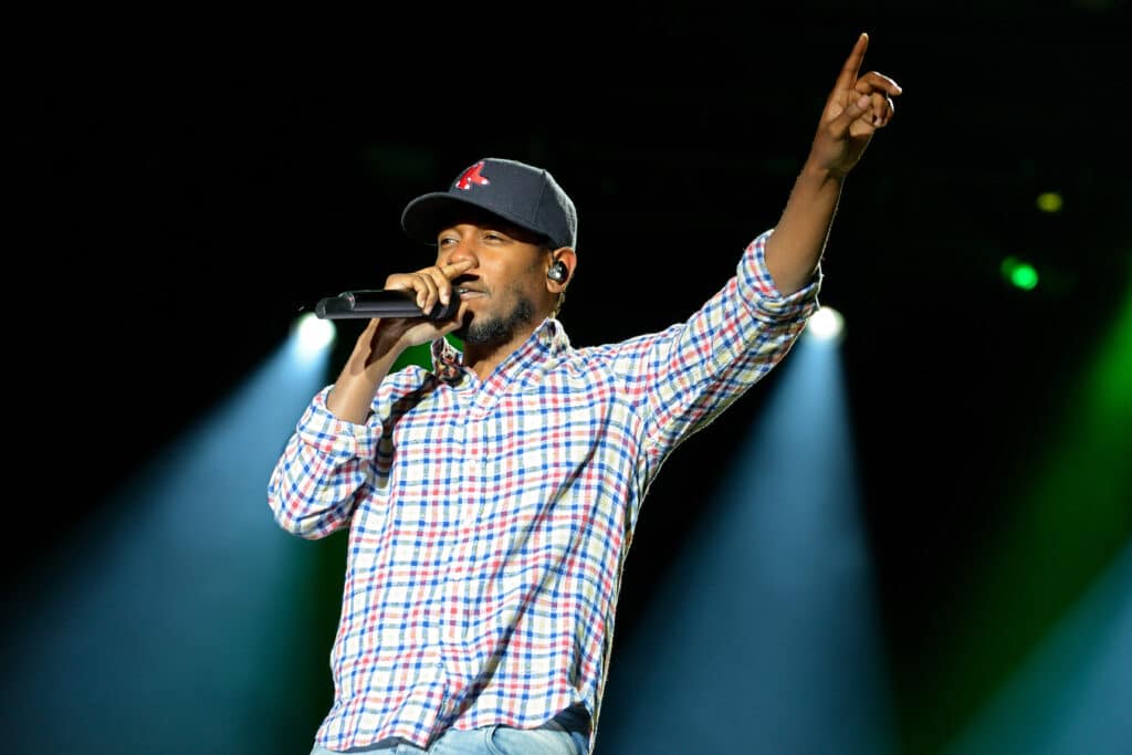 Kendrick Lamar is performing on stage. With one hand he is holding a microphone up to his mouth. With the other, he is pointing up.